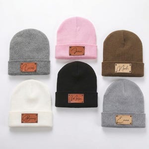 Peut inclure: Six bonnets en tricot de couleurs variées : gris, rose, noir, blanc, marron et gris clair. Chaque bonnet est orné d'un patch rectangulaire en cuir avec un nom. Les noms sont Lucas, Jamie, Mason, Benny, Noah et Emma.