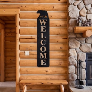 Puede incluir: Un letrero de pared vertical de metal negro que presenta la silueta de un oso en la parte superior y la palabra WELCOME impresa en letras mayúsculas grandes y claras. El letrero rectangular tiene esquinas redondeadas y está montado en una pared de troncos.