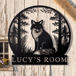 May include: Black metal wall art featuring a fox in a forest scene, framed by a circular design. The fox has a white chest and is surrounded by trees, ferns, and grass. The text "LUCY'S ROOM" is displayed below the circular design.