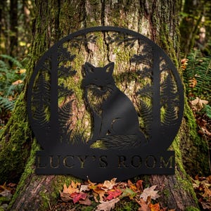 May include: A black metal sign featuring a fox in a forest scene. The fox is centered within a circular frame with trees and ferns. The text "LUCY'S ROOM" is displayed at the bottom of the sign. The sign is mounted on a tree trunk.