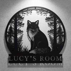 May include: Black metal wall art featuring a fox in a forest scene. The fox is centered within a circular frame with trees, ferns, and foliage. The text "LUCY'S ROOM" is displayed below the circular frame.