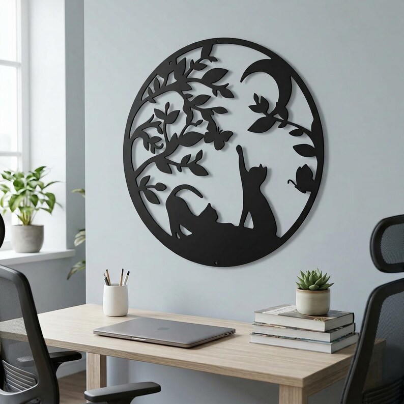 May include: Black metal wall art featuring a circular design with two cats, a tree, a crescent moon, and butterflies. The cats are in silhouette, one reaching up. The art is mounted on a light blue wall.