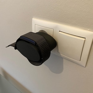 Könnte beinhalten: Ein schwarzer und grauer Fitnesstracker mit einem dunkelgrauen gewebten Armband ist in eine weiße Steckdose an einer weißen Wand eingesteckt. Der Bildschirm des Trackers ist dunkel und das Armband ist um die Steckdose gewickelt.