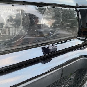 Pode incluir: Um conjunto de faróis de carro pretos com um dispositivo preto retangular de dois botões montado no para-choques. Os faróis são transparentes e o carro é preto.