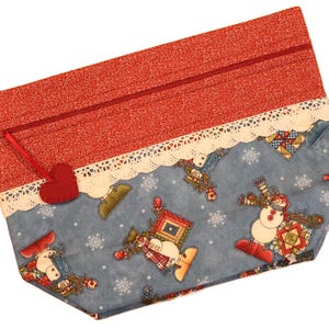 May include: A handmade zippered pouch with a festive winter theme. The top section is red with a small white pattern, and the bottom features a blue fabric with snowmen and snowflakes. A red heart charm hangs from the zipper.