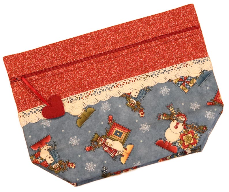 May include: A handmade zippered pouch with a festive winter theme. The top section is red with a small white pattern, and the bottom features a blue fabric with snowmen and snowflakes. A red heart charm hangs from the zipper.