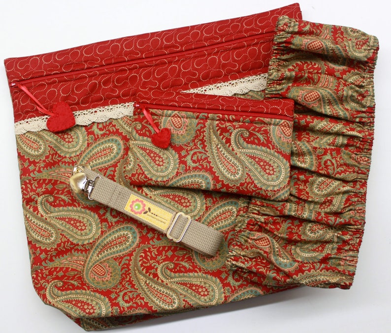 May include: Two red and gold paisley patterned zippered pouches with white lace trim and red pom-poms. The larger pouch has a tan strap with a silver clip.