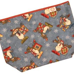 May include: A blue fabric pouch with a repeating pattern of snowmen, snowflakes, and patchwork quilt designs. The pouch has a red glitter trim and a small tag. The snowmen are wearing hats, scarves, and mittens.