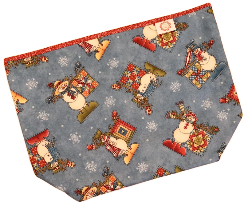 May include: A blue fabric pouch with a repeating pattern of snowmen, snowflakes, and patchwork quilt designs. The pouch has a red glitter trim and a small tag. The snowmen are wearing hats, scarves, and mittens.