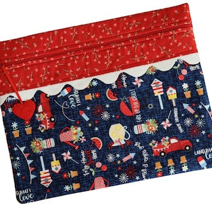 May include: A red, white, and blue fabric zipper pouch with a patriotic theme. The pouch features a variety of designs, including fireworks, stars, and the words "Land That I Love".