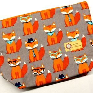 Pode incluir: Uma bolsa de tecido cinzento com fecho de correr e um padr&atilde;o repetido de raposas laranja que usam &oacute;culos, chap&eacute;us e gravatas. A bolsa tem uma etiqueta que diz "Facebook & Etsy SewMuchCutest.com".