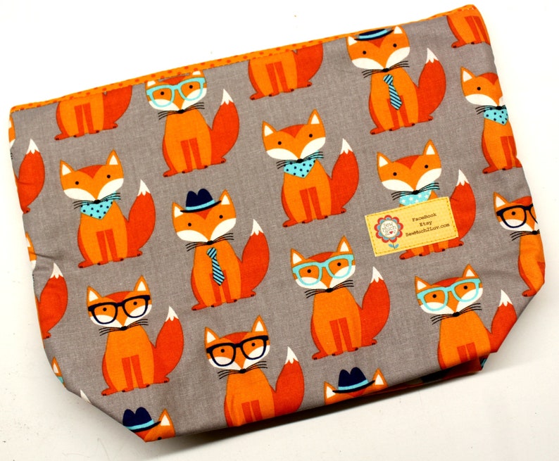 Pode incluir: Uma bolsa de tecido cinzento com fecho de correr e um padr&atilde;o repetido de raposas laranja que usam &oacute;culos, chap&eacute;us e gravatas. A bolsa tem uma etiqueta que diz "Facebook & Etsy SewMuchCutest.com".