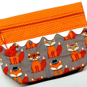 Pode incluir: Uma bolsa de z&iacute;per cinza com uma borda branca festonada e um padr&atilde;o repetido de raposas laranja usando &oacute;culos e chap&eacute;us. A bolsa tem um z&iacute;per vermelho e um pequeno amuleto de cora&ccedil;&atilde;o laranja.