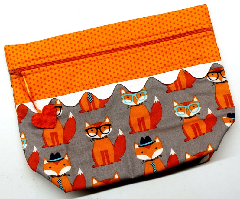 Pode incluir: Uma bolsa de z&iacute;per cinza com uma borda branca festonada e um padr&atilde;o repetido de raposas laranja usando &oacute;culos e chap&eacute;us. A bolsa tem um z&iacute;per vermelho e um pequeno amuleto de cora&ccedil;&atilde;o laranja.