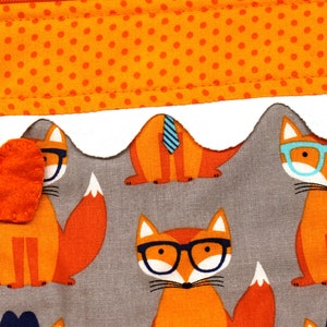 Pode incluir: Uma bolsa de tecido cinza com uma borda branca festoneada e um fecho de correr. A bolsa &eacute; decorada com um padr&atilde;o repetido de raposas laranja que usam &oacute;culos. Um pequeno cora&ccedil;&atilde;o de feltro laranja est&aacute; preso ao puxador do fecho de correr.