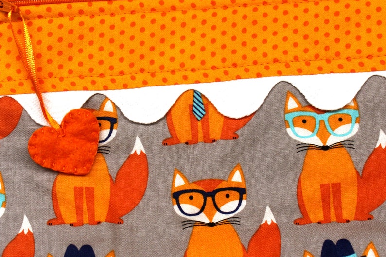 Pode incluir: Uma bolsa de tecido cinza com uma borda branca festoneada e um fecho de correr. A bolsa &eacute; decorada com um padr&atilde;o repetido de raposas laranja que usam &oacute;culos. Um pequeno cora&ccedil;&atilde;o de feltro laranja est&aacute; preso ao puxador do fecho de correr.