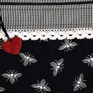 May include: A black fabric bag with a white and black checkered pattern and a white lace trim. A red felt heart hangs from the top of the bag. The bag is decorated with a repeating pattern of white bees.