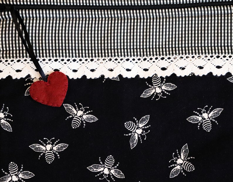 May include: A black fabric bag with a white and black checkered pattern and a white lace trim. A red felt heart hangs from the top of the bag. The bag is decorated with a repeating pattern of white bees.