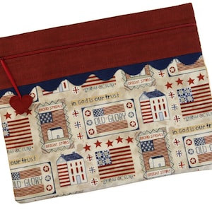 May include: A red, white, and blue fabric zippered pouch with a heart-shaped charm. The fabric features a repeating pattern of American flags, houses, and the words "In God is our trust", "Old Glory", "Grand Old Flag", "Bright Stars", and "Broad Stripes".