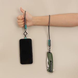 Puede incluir: Un cordón gris con detalles turquesa y un teléfono negro adjunto. Un segundo cordón gris con detalles turquesa está sujeto a una navaja plegable verde. Los artículos cuelgan sobre un fondo neutro.