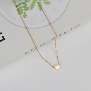 Puede incluir: Un delicado collar dorado con un pequeño colgante en forma de corazón. La cadena es fina y el corazón es liso y pulido. El collar se muestra sobre una superficie blanca, creando una estética simple y elegante.