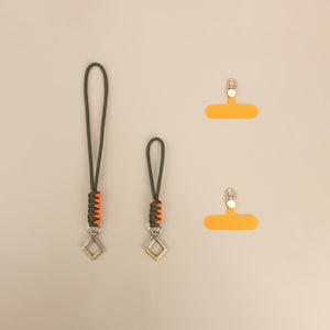Puede incluir: Dos colgantes para teléfono verde oliva y naranja con cierres en forma de diamante. También se muestran dos pestañas de agarre para teléfono naranja con cierres plateados. Los colgantes tienen un diseño tejido naranja y verde.