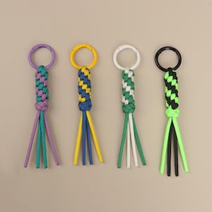 Puede incluir: Cuatro llaveros coloridos con cordón trenzado y anillos metálicos. Los llaveros presentan cordones morados, amarillos, blancos, verdes, negros y azules. Cada llavero tiene una combinación de colores diferente.