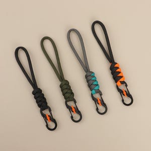 Pode incluir: Quatro chaveiros de paracord nas cores preto, verde-oliva, cinza e preto com detalhes em laranja. Cada chaveiro possui uma alça, uma seção tecida e um mosquetão preto com detalhes em laranja. Em um fundo neutro.