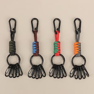 May include: Four keychains with black carabiner clips and key rings. Each keychain has a different colored woven cord: green and black, blue and brown, red and green, and gray and orange. The key rings each hold six small black clips.