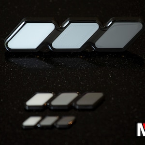 May include: A set of three black-framed, rhomboid-shaped decorative pieces arranged in a row, with three smaller sets of identical pieces positioned below in decreasing sizes, each featuring a light-coloured, reflective centre panel.