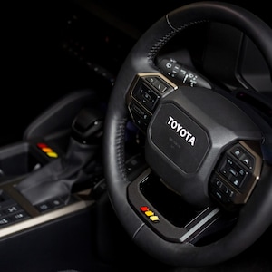 May include: A black Toyota steering wheel with gold accents. The steering wheel has the Toyota logo in white. The image also shows the gear shift and console of the vehicle. The steering wheel has buttons and a display.