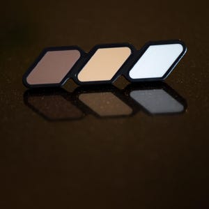 Puede incluir: Un emblema negro con tres elementos en forma de rombo desplazados en marrón, beige y blanco. El emblema tiene un acabado brillante sobre un fondo oscuro. La esquina inferior derecha muestra el texto "MX DESIGN" en un rectángulo blanco.