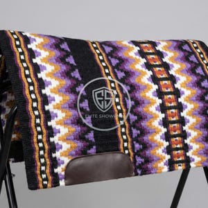 May include: A colorful horse saddle pad with a geometric pattern in black, purple, white, and orange. The pad is displayed on a black metal stand, showcasing its design and texture. The pad has a brown leather accent.