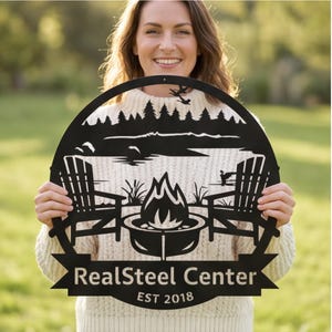 May include: Black metal sign with a lakeside scene, featuring Adirondack chairs, a fire pit, and trees. The sign has the text "RealSteel Center EST 2018" in a banner across the bottom. The sign is held by a person.