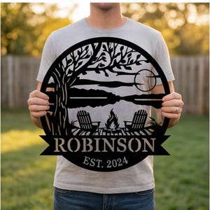 May include: A circular black metal wall sign featuring a laser-cut outdoor scene with a tree, two chairs, a campfire, and a moon. The sign includes a banner with the name ROBINSON and the text EST. 2024 printed in a bold, sans-serif font.