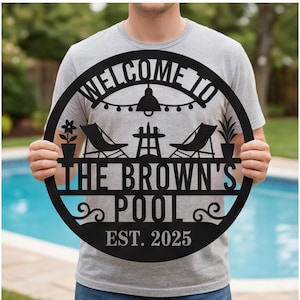 May include: A circular black metal sign featuring a cutout design of two lounge chairs, a small table, a potted plant, and a flower. The text reads WELCOME TO THE BROWN'S POOL EST. 2025, with a string of lights depicted above the furniture.