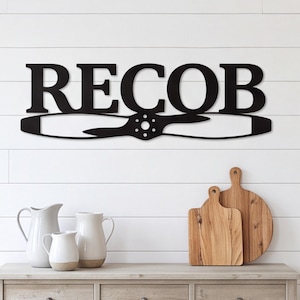 以下が含まれることがあります： 太字のセリフ体で「RECOB」という文字がデザインされた黒い金属製のウォールサイン。そのすぐ下には飛行機のプロペラのシルエットが配置されています。サインは、木製の表面の上にある白い水平の羽目板の壁に取り付けられています。