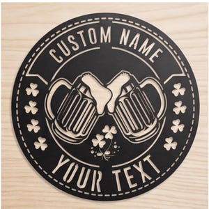 Könnte beinhalten: Runder Metallschild in Schwarz mit den weißen Schriftzügen "CUSTOM NAME" und "YOUR TEXT". Das Design zeigt zwei Bierkrüge, die anstoßen, mit überschäumendem Schaum. Kleeblattmotive sind ebenfalls enthalten.