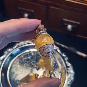 Puede incluir: Una botella de poción dorada con tapón de corcho y base plateada. La botella está llena de un líquido dorado brillante y tiene una etiqueta que dice "Felix Felicis".