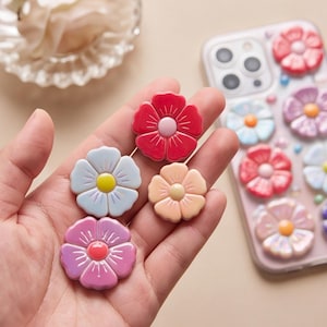 May include: A collection of small, flat, flower-shaped charms in various colors including red, light blue, pink, and peach. Each charm features five rounded petals and a central circular accent. A clear smartphone case decorated with similar flower charms.