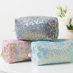 Puede incluir: Tres neceseres con lentejuelas en colores pastel: azul claro, rosa iridiscente y plateado. Cada bolsa rectangular tiene cierre de cremallera y un dije de perla. Flores blancas en un jarrón en el fondo.
