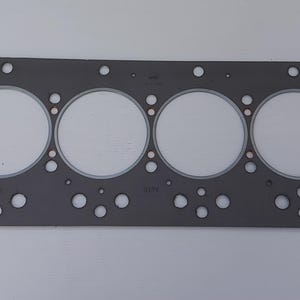 Puede incluir: Una junta de culata gris con seis grandes aberturas circulares y numerosos agujeros m&aacute;s peque&ntilde;os. La junta es rectangular con esquinas redondeadas y est&aacute; dise&ntilde;ada para un motor. El material parece ser un compuesto de papel y metal.