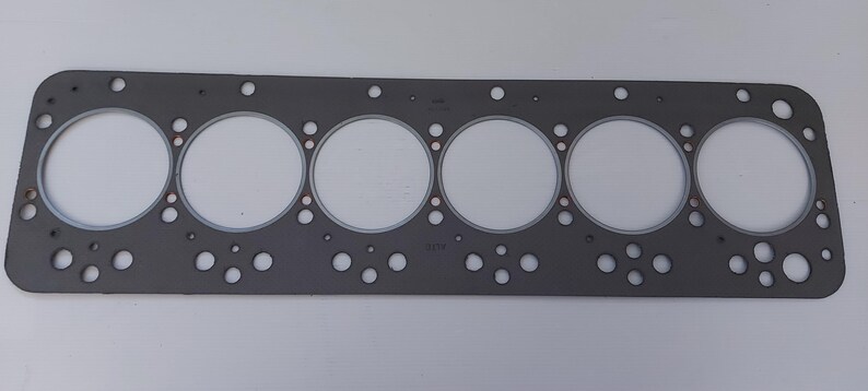 Puede incluir: Una junta de culata gris con seis grandes aberturas circulares y numerosos agujeros m&aacute;s peque&ntilde;os. La junta es rectangular con esquinas redondeadas y est&aacute; dise&ntilde;ada para un motor. El material parece ser un compuesto de papel y metal.