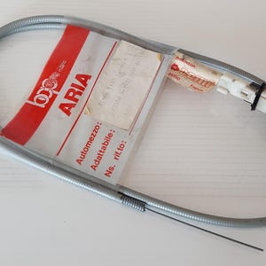 May include: A silver-colored cable with a black knob and a white plastic component. The cable is coiled and has a label with the word "ARIA" in red and white. The cable is likely an automotive part.