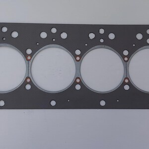 Puede incluir: Una junta de culata de motor gris con seis grandes aberturas circulares y numerosos agujeros m&aacute;s peque&ntilde;os. La junta tiene forma rectangular con esquinas redondeadas y detalles en color cobre alrededor de las aberturas circulares. El fondo es blanco liso.