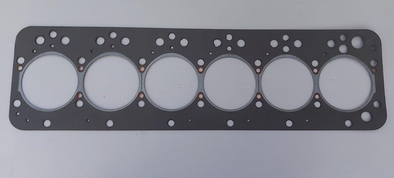 Puede incluir: Una junta de culata de motor gris con seis grandes aberturas circulares y numerosos agujeros m&aacute;s peque&ntilde;os. La junta tiene forma rectangular con esquinas redondeadas y detalles en color cobre alrededor de las aberturas circulares. El fondo es blanco liso.