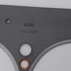 Puede incluir: Una junta gris con m&uacute;ltiples agujeros circulares y un borde de metal plateado. Los textos "SECTOR" y "specs50" est&aacute;n impresos en la junta. Un anillo de color cobre es visible en la parte inferior.