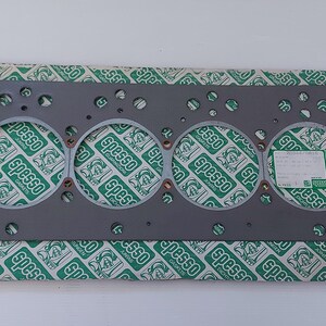 Puede incluir: Una junta de culata gris con seis grandes aberturas circulares y numerosos agujeros m&aacute;s peque&ntilde;os. La junta est&aacute; sobre papel estampado verde y blanco. Una peque&ntilde;a etiqueta est&aacute; adherida al papel.