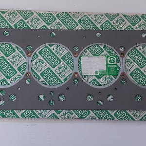 Puede incluir: Una junta de culata gris con seis grandes aberturas circulares y numerosos agujeros m&aacute;s peque&ntilde;os. La junta est&aacute; sobre un papel estampado verde y blanco con la palabra "spesso" repetida. Se adjunta una peque&ntilde;a etiqueta.