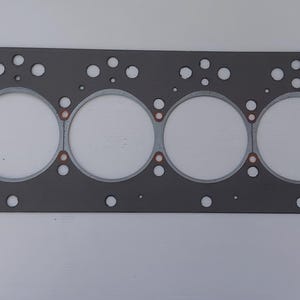 Puede incluir: Una junta de culata rectangular gris con seis grandes aberturas circulares y numerosos agujeros m&aacute;s peque&ntilde;os. La junta tiene un borde gris claro alrededor de las grandes aberturas y detalles en color cobre.